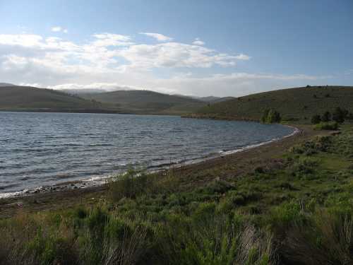 A serene lake surrounded by rolling hills and greenery under a partly cloudy sky.