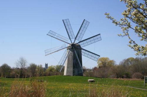A traditional windmill with large sails stands in a green field, surrounded by trees and a clear blue sky.