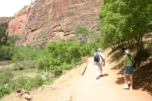 Two hikers walking on a dirt path surrounded by lush greenery and towering red rock formations.