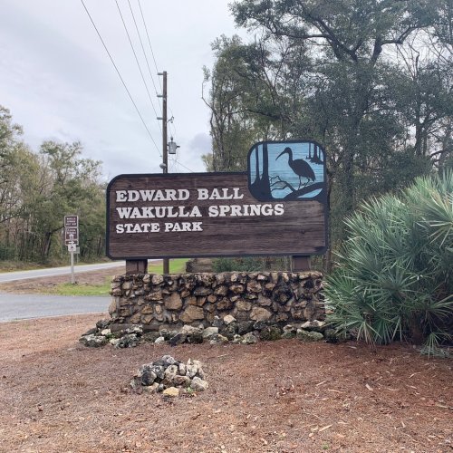 Sign for Edward Ball Wakulla Springs State Park, featuring a bird illustration and surrounded by trees and gravel.