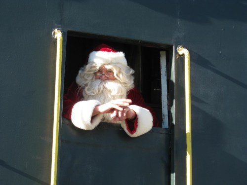 Santa Claus with a white beard and red suit leans out of a window, smiling and waving.