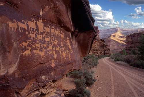 Petroglyphs on a rock face in a canyon, with a dirt road and dramatic clouds in the background.