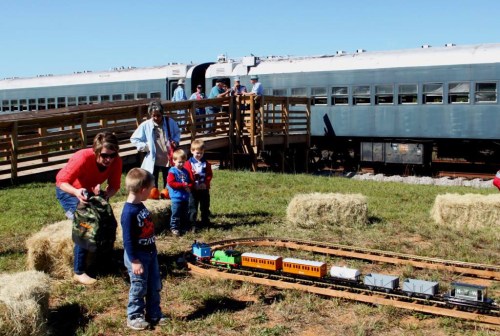 Children play with a toy train on a track, while adults watch near vintage train cars and hay bales in a sunny field.