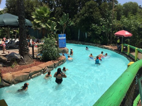 A sunny water park scene with people swimming in a lazy river surrounded by lush greenery and lounge chairs.