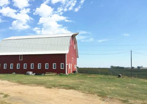 A red barn with a metal roof stands in a grassy field under a blue sky with scattered clouds.