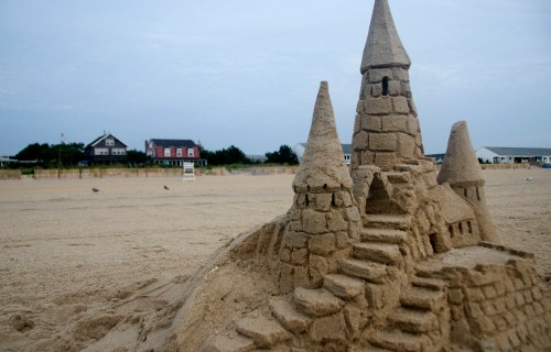 A detailed sandcastle with towers and stairs, set on a beach with a cloudy sky and distant buildings in the background.