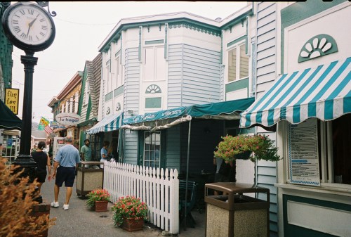 A quaint street scene featuring colorful buildings, striped awnings, and a clock, with people walking and flowers in planters.