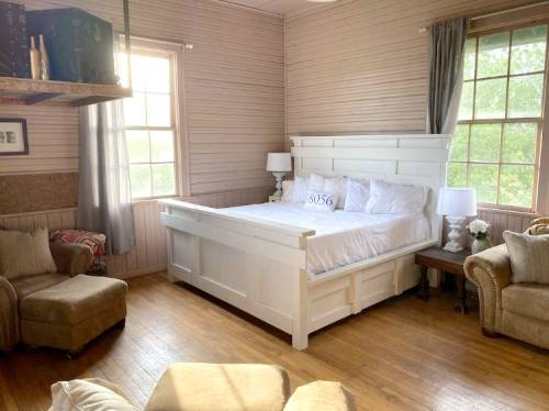 Cozy bedroom with a white bed, two lamps, and armchairs, featuring large windows and warm wooden floors.