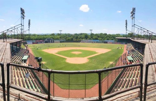 A panoramic view of a baseball field with empty stands and players on the field under a clear blue sky.