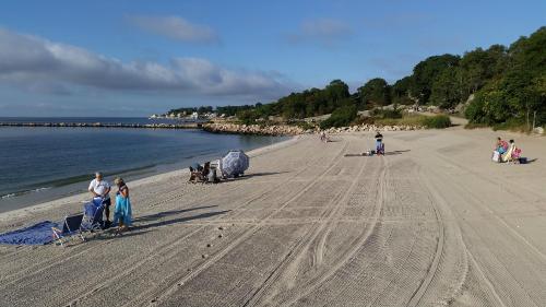A sandy beach with a calm sea, people relaxing under umbrellas, and a rocky jetty in the background.