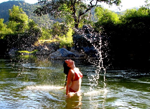 A person in a red bikini splashes water while standing in a river, surrounded by trees and sunlight.