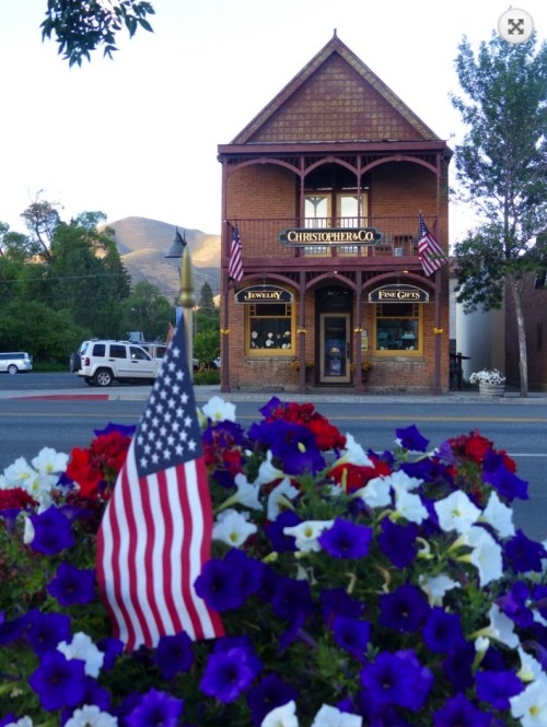 Historic brick building with American flags, featuring a jewelry store and colorful flower beds in the foreground.