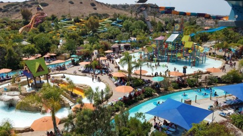 A vibrant water park scene with pools, slides, and colorful umbrellas, filled with people enjoying the sunny day.