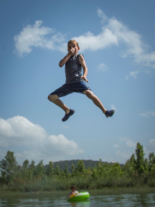 A boy jumps joyfully into a lake, with a second child in a float nearby, under a blue sky with fluffy clouds.