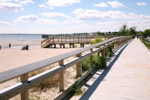 A sandy beach with a wooden boardwalk, people walking, and a pier in the distance under a partly cloudy sky.
