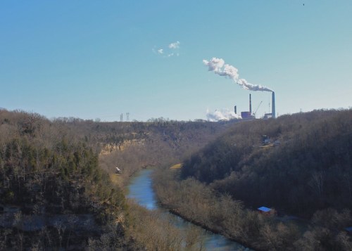 A river flows through a valley with a power plant emitting smoke in the background under a clear blue sky.