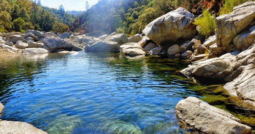 A serene river scene with clear water, surrounded by large rocks and lush greenery under a bright blue sky.