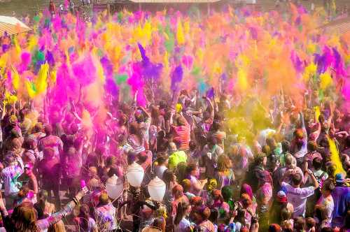 A vibrant crowd celebrating with colorful powders, creating a cloud of pink, purple, and yellow hues in the air.