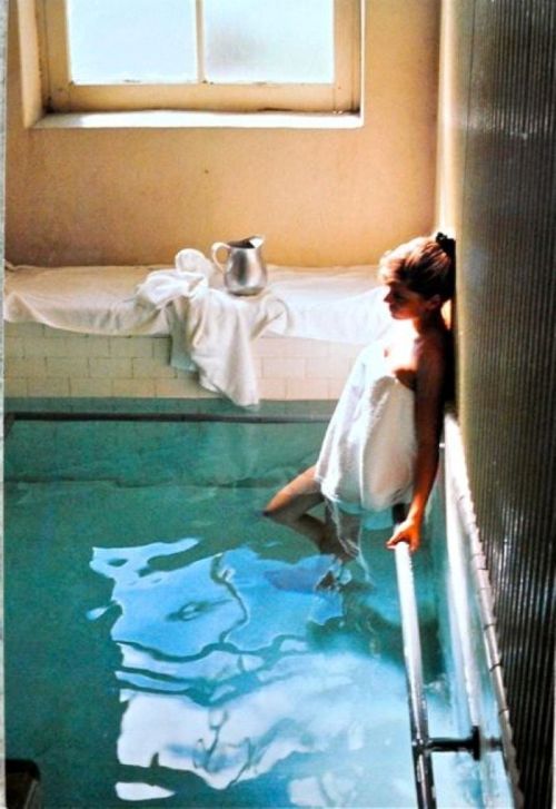 A girl in a white towel leans against a pool's edge, sunlight streaming through a window.