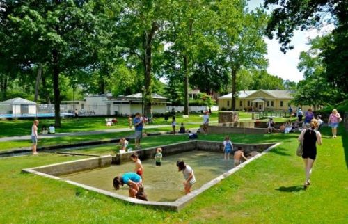 A sunny park scene with children playing in a shallow pool, surrounded by trees and people enjoying the outdoors.