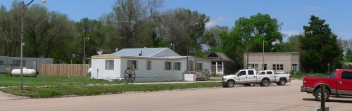 A small rural scene featuring a white mobile home, a few vehicles, and green trees under a clear blue sky.