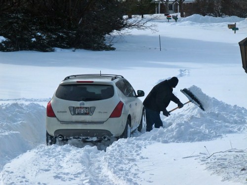 A person shovels snow beside a car stuck in a snowy driveway, with deep snow covering the ground.