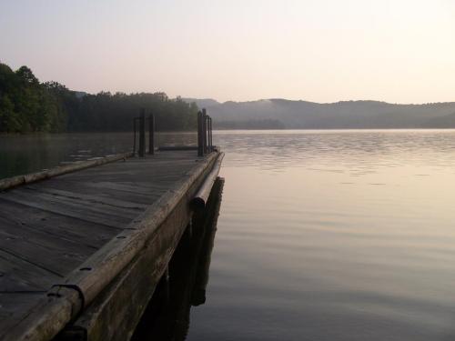 A serene wooden dock extends into a calm lake, surrounded by misty mountains and trees at dawn.