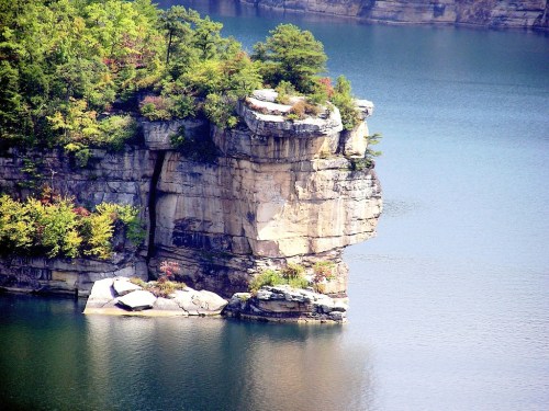A rocky cliff with greenery overlooks a calm, blue lake, reflecting the surrounding landscape.