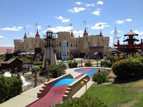 A colorful mini-golf course featuring a castle, lighthouse, and various themed structures under a blue sky.