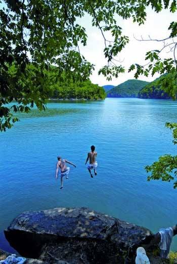 Two people jumping into a serene blue lake surrounded by green trees and mountains on a sunny day.