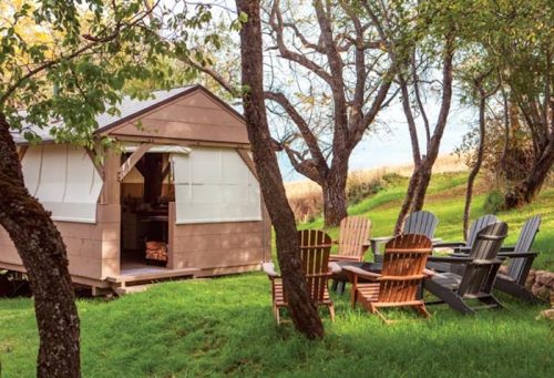 A cozy cabin surrounded by trees, with colorful chairs arranged around a fire pit on a grassy area.