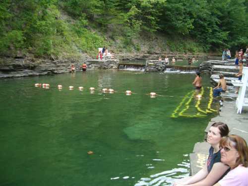 People relax by a clear, green swimming area with rocky edges and a gentle waterfall in a lush, wooded setting.