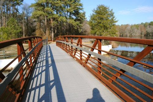 A metal bridge spans a calm waterway, surrounded by trees and a clear blue sky, casting long shadows on the walkway.