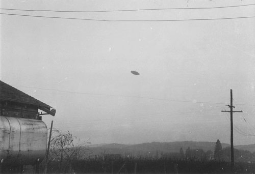 A black and white photo of a UFO hovering above a rural landscape with a house and power lines in the foreground.