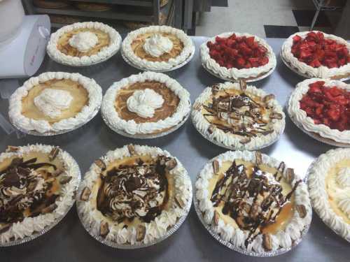 A variety of pies with whipped cream and fruit toppings arranged on a table in a bakery.