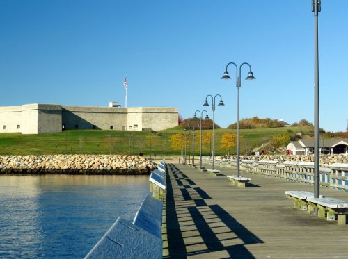 A wooden pier extends over calm water, with a historic fort and colorful trees in the background under a clear blue sky.