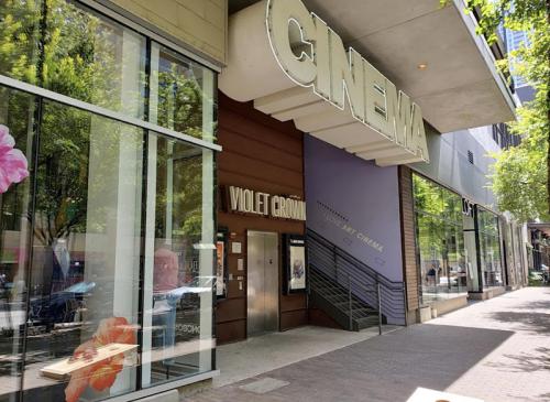 Entrance of a cinema building with large "CINEMA" sign and "Violet Crown" marquee, surrounded by trees and urban setting.