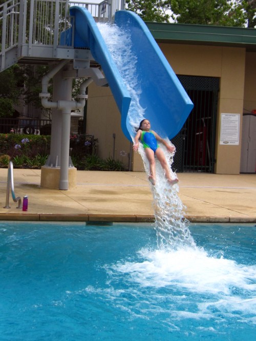 A child slides down a blue water slide, splashing into a pool below on a sunny day.