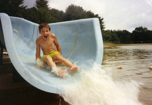 A young boy joyfully slides down a water slide into a lake, splashing water around him on a sunny day.