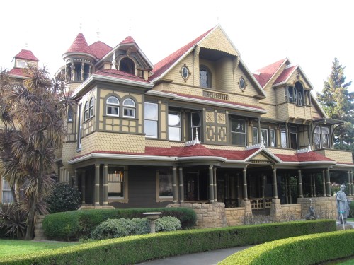 A large, ornate Victorian-style mansion with multiple gables and a wraparound porch, surrounded by lush greenery.