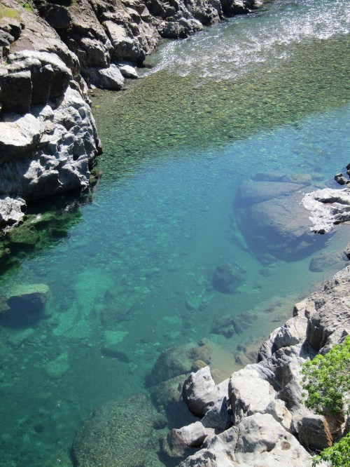A clear, turquoise river flows between rocky banks, revealing stones beneath the water's surface.