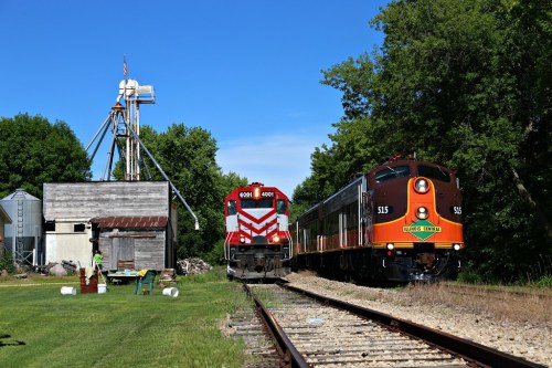 Two vintage trains parked beside a rustic building and greenery under a clear blue sky.