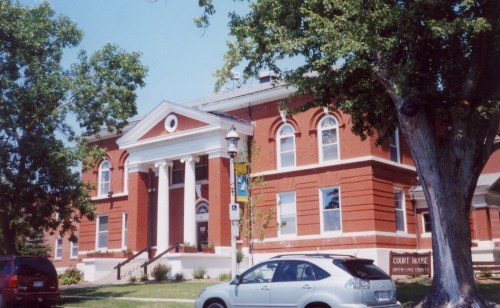 Historic brick courthouse with white columns, surrounded by trees and parked cars. Clear blue sky in the background.
