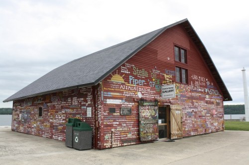 A red barn-like building covered in colorful signs and text, with a door open to a gallery, near a body of water.