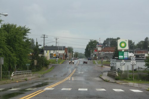 A rainy street scene with a gas station, shops, and traffic signs, lined with trees and buildings in the background.
