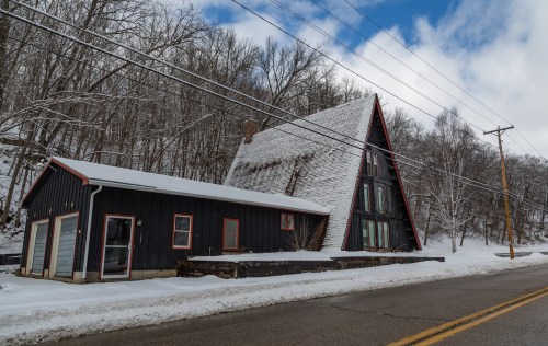 A snowy landscape featuring a unique A-frame house and a smaller building, surrounded by bare trees and a cloudy sky.
