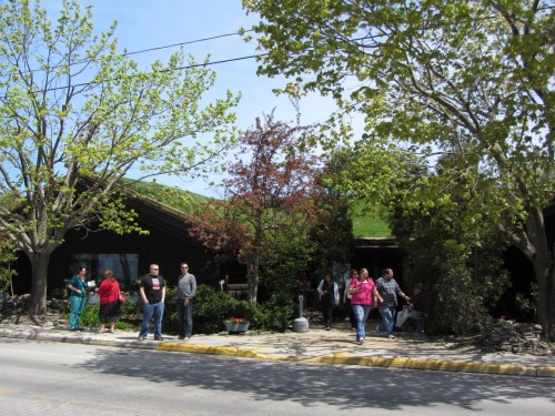 A group of people stands outside a rustic building surrounded by trees on a sunny day.