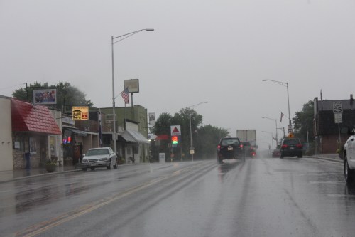 A rainy street scene with cars driving, shops lining the road, and an American flag visible in the background.