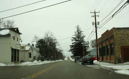 A snowy street scene with houses and businesses, power lines, and trees under a gray sky.