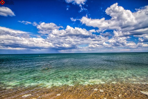 A serene seascape featuring clear blue water, a sandy shore, and a sky filled with fluffy clouds.
